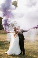 Braut und Bräutigam spielen mit farbig Rauch im lila Hände, Umarmungen und Küsse. Rauch Bomben beim ein Hochzeit. foto