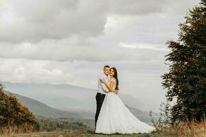 ein Hochzeit Paar zärtlich Umarmungen im das Berge. das Bräutigam Umarmungen das Braut durch das Schultern. ein Herrlich und großartig Braut Kleid. foto