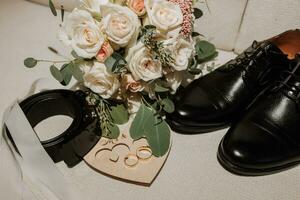 Einzelheiten von das Bräutigam sind vereinbart worden im ein Komposition. schwarz Schuhe, Hochzeit Strauß, Gold Hochzeit Ringe auf ein hölzern Stand, schwarz Herren Gürtel foto