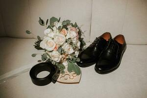 Einzelheiten von das Bräutigam sind vereinbart worden im ein Komposition. schwarz Schuhe, Hochzeit Strauß, Gold Hochzeit Ringe auf ein hölzern Stand, schwarz Herren Gürtel foto