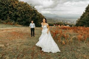 Hochzeit Paar Gehen im das Berge. das Braut Spaziergänge zu das Bräutigam suchen in das Kamera. das Braut Kleid entwickelt im das Wind. wunderbar Herbst Wetter foto