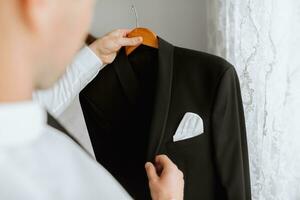 ein jung Mann hält seine schwarz Jacke auf ein Aufhänger im seine Hände. das Bräutigam ist vorbereiten zum das Hochzeit Zeremonie. detailliert Nahansicht Foto von Hände