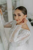 ein schön Braut im ein Hochzeit Kleid mit geschlossen Schultern steht durch ein Spiegel im ihr Zimmer, sieht aus in das Linse. foto