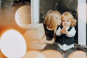 fröhlich Weihnachten und glücklich Feiertage. heiter Mädchen und Junge sind Sitzung auf das Fensterbrett in der Nähe von das Weihnachten Baum mit Weihnachten die Geschenke. Kinder sind haben Spaß. liebend Familie mit Geschenke im das Zimmer. foto