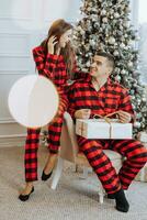 jung schön Familie Paar im identisch rot Pyjama in der Nähe von das Weihnachten Baum jubeln und Austausch Weihnachten Geschenke im ihr Hände. Neu Jahre Ferien und Geschenke unter das Weihnachten Baum foto