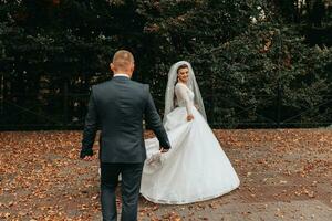 Hochzeit Porträt. ein Brünette Braut im ein lange Kleid hält ihr Strauß von Rosen, Pfingstrosen und Grün, das Bräutigam im ein klassisch passen Spaziergänge hinter ihr auf das Brücke. foto