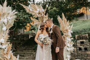 Bräutigam und Braut Kuss. Jungvermählten mit ein Hochzeit Strauß Stehen beim ein Hochzeit Zeremonie unter ein Bogen dekoriert mit Blumen und getrocknet Blumen draußen. foto