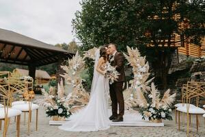 Bräutigam und Braut Kuss. Jungvermählten mit ein Hochzeit Strauß Stehen beim ein Hochzeit Zeremonie unter ein Bogen dekoriert mit Blumen und getrocknet Blumen draußen. foto
