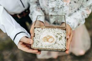 Hochzeit Ringe von das Braut und Bräutigam im ein Glas Box auf ein Pfingstrose Blume. Kinder- Hände halt Hochzeit Ringe. Nahansicht foto