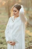 ein schwanger Frau im ein Weiß Kleid auf ein dunkel Hintergrund posiert im das Park. glücklich und sorglos Schwangerschaft. Herbst Park. glücklich Frau genießen ihr Schwangerschaft. foto