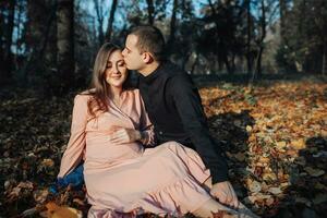 ein Mann berührt seine schwanger Ehefrauen Bauch mit seine Hand während ruhen im ein Herbst Wald auf ein vergilbt Blatt. Unterstützung von das Mann während Schwangerschaft foto