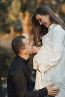 jung schwanger Frau im ein Weiß Shirt. das Mann stellen seine Hände auf seine Ehefrauen Bauch und beugte sich vor gegen seine Ohr. foto