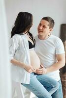 modern schön schwanger Frau im Jeans und Hemd mit Mann umarmen ihr Bauch mit Hände im schön Sonnenlicht. Konzept von Schwangerschaft, Mutterschaft, Vorbereitung und warten. foto