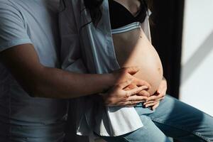 modern schön schwanger Frau im Jeans und Hemd mit Mann umarmen ihr Bauch mit Hände im schön Sonnenlicht. Konzept von Schwangerschaft, Mutterschaft, Vorbereitung und warten. foto