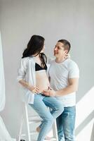 modern schön schwanger Frau im Jeans und Hemd mit Mann umarmen ihr Bauch mit Hände im schön Sonnenlicht. Konzept von Schwangerschaft, Mutterschaft, Vorbereitung und warten. foto