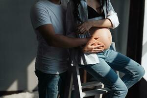 modern schön schwanger Frau im Jeans und Hemd mit Mann umarmen ihr Bauch mit Hände im schön Sonnenlicht. Konzept von Schwangerschaft, Mutterschaft, Vorbereitung und warten. foto