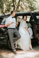 Hochzeit Porträt, Foto von ein stilvoll Bräutigam im ein Weiß Hemd und Bogen Krawatte und ein Brünette Braut mit ein Strauß von Blumen in der Nähe von ein schwarz Wagen.