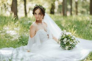 ein schön Braut im ein Weiß Kleid mit ein Kranz von frisch Blumen ist Sitzung auf das Grün Gras foto