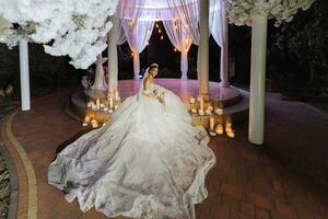 ein jung schön Braut im ein Hochzeit Kleid mit öffnen Schultern und ein Krone auf ihr Kopf ist Sitzung auf das Treppe im das Abend Beleuchtung nach das Hochzeit Zeremonie. foto