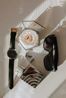 Einzelheiten von das Bräutigam auf ein Weiß Tisch. schwarz Gläser, ein Armbanduhr, Gold Hochzeit Ringe, Herren Parfums, Manschettenknöpfe foto