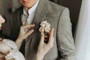 Seite Aussicht von Braut mit Hochzeit Frisur im Hochzeit Kleid halten Boutonniere auf gut aussehend Bräutigam grau Anzug, Paar haben Morgen Vorbereitung zum Engagement Tag. foto