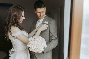 Seite Aussicht von Braut mit Hochzeit Frisur im Hochzeit Kleid halten Boutonniere auf gut aussehend Bräutigam grau Anzug, Paar haben Morgen Vorbereitung zum Engagement Tag. foto