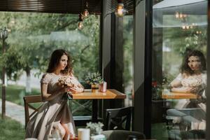ein jung stilvoll Frau im ein Beige Kleid mit ein Strauß von Blumen im ihr Hände ist Sitzung im ein schön Licht im ein Cafe im Spanien foto