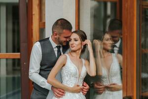 stilvoll Braut und Bräutigam im ein Hochzeit Kleid mit ein lange Zug und ein Schleier sind Stehen in der Nähe von ein Glas Vitrine. Vorderseite Foto