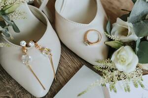Weiß Hochzeit Schuhe und Einzelheiten von das Braut. Hochzeit Ring mit Diamant und Gold Armband mit Kristalle, auf hölzern Hintergrund. Blumen und Grün. Hochzeit Thema foto