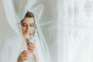 das Braut ist gekleidet im ein elegant Kleid, bedeckt mit ein Schleier, posieren und halten ein Ansteckblume. Hochzeit Foto, Morgen von das Braut foto