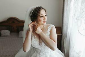 Porträt von das Braut. das Braut ist gekleidet im ein elegant Kleid, posieren durch das Fenster tragen Ohrringe. Tiara und Schmuck. öffnen Büste. Morgen von das Braut foto