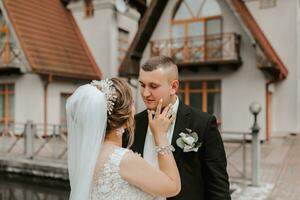 ein Brünette Braut im ein lange Kleid und ein Bräutigam im ein klassisch passen Stand Umarmen auf ein Brücke in der Nähe von ein See mit ein Schloss im das Hintergrund. foto