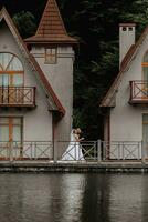 ein Brünette Braut im ein lange Kleid und ein Bräutigam im ein klassisch passen sind Gehen auf ein Brücke in der Nähe von ein See gegen das Hintergrund von ein Schloss. Betrachtung im das Wasser von das Schloss. symmetrisch Anzeige foto