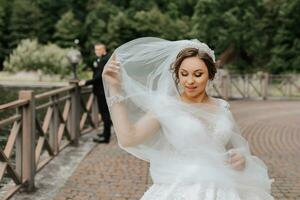 Hochzeit Porträt. das Braut im ein Weiß Kleid auf das Brücke mit ein fließend Schleier, das Bräutigam ist Stehen hinter ihr. aufrichtig lächeln. Wind und Schleier. Diadem. foto