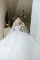 Vorbereitung zum das Hochzeit. schön jung Braut im Weiß luxuriös Hochzeit Kleid, Tiara auf Kopf, lange Schleier im königlich Hotel Zimmer. Luxus lächelnd Modell- foto