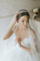 Vorbereitung zum das Hochzeit. schön jung Braut im Weiß luxuriös Hochzeit Kleid, Tiara auf Kopf, lange Schleier im königlich Hotel Zimmer. Luxus lächelnd Modell- foto