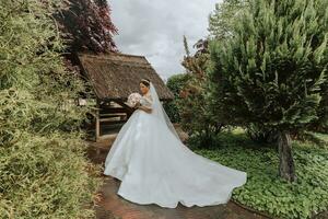 Rückseite Aussicht Porträt von schön Braut im Weiß Hochzeit Kleid mit lange Zug mit modern Frisur und Schleier Gehen im Garten. Hochzeit Konzept foto