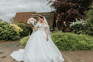 Porträt von ein schön Braut im ein Weiß Hochzeit Kleid mit ein lange Zug mit ein modern Frisur und ein Schleier Gehen im das Garten. Hochzeit Konzept foto