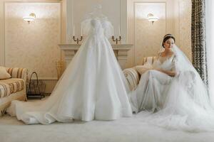 ein schön Mädchen mit ein Hochzeit Frisur im ein transparent Kleid ist vorbereiten zum ein Hochzeit im ein Hotel mit ein königlich Innere. Hochzeit Kleid auf ein Mannequin im natürlich Licht foto