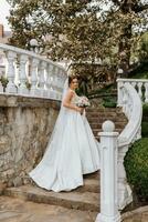 ein schön Brünette Braut im ein Weiß lange Kleid mit ein Strauß von Blumen steht auf das Treppe gegen das Hintergrund von hoch Bäume, draußen im das Park. foto