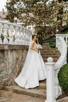 ein schön Brünette Braut im ein Weiß lange Kleid mit ein Strauß von Blumen steht auf das Treppe gegen das Hintergrund von hoch Bäume, draußen im das Park. foto