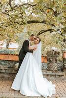 modisch Bräutigam und süß Brünette Braut im Weiß Kleid mit Fachmann bilden und Strauß von Blumen sind umarmen, Lachen im Park, Garten, Wald draußen. Hochzeit Fotografie foto