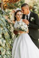 ein stilvoll Bräutigam im ein schwarz passen und ein süß Braut im ein Weiß Kleid mit ein lange Schleier sind umarmen im ein Park in der Nähe von ein alt Haus. Hochzeit Porträt von lächelnd und glücklich Jungvermählten. foto