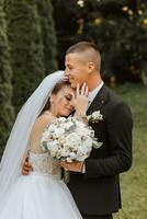 ein stilvoll Bräutigam im ein schwarz passen und ein süß Braut im ein Weiß Kleid mit ein lange Schleier sind umarmen im ein Park. Hochzeit Porträt von lächelnd und glücklich Jungvermählten. foto