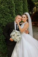 ein stilvoll Bräutigam im ein schwarz passen und ein süß Braut im ein Weiß Kleid mit ein lange Schleier sind umarmen im ein Park. Hochzeit Porträt von lächelnd und glücklich Jungvermählten. foto