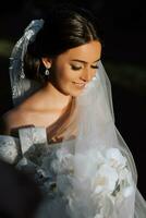 zärtlich Porträt von ein glücklich Braut mit ein Strauß von Orchideen im ihr Hände, draußen. warm Sommer- Zeit. ein lange Schleier, ein luxuriös Weiß Kleid mit ein Zug. foto