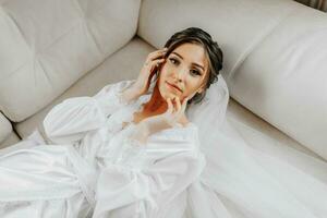 ein schön Braut ist Lügen auf ein modern Sofa im ein Weiß Kleid. Hochzeit Frisur, Fachmann bilden. romantisch Braut posieren foto