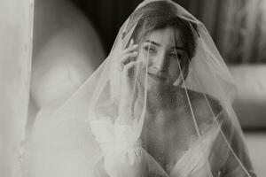 ein atemberaubend jung Braut posiert durch das Fenster halten ihr Schleier im ihr Hände. ein schön Frau von kaukasisch Aussehen im ein elegant Weiß Kleid und luxuriös Frisur. Hochzeit bilden foto