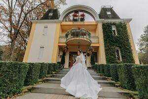 Herrlich Braut im Hochzeit Kleid mit lange Zug und Schleier draußen. das Braut gegen das Hintergrund von ein teuer Hotel foto