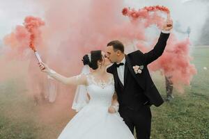 schön Jungvermählten Stand im ein Feld gegen das Hintergrund von Berge und halt mehrfarbig Rauch Bomben im ihr Hände. Hochzeit Fotografie. foto